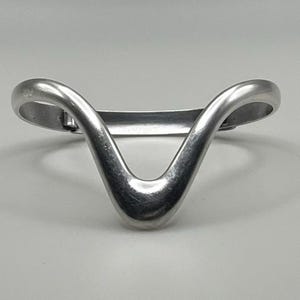 May include: A silver-colored, V-shaped bracelet. The bracelet has a smooth, curved design with rounded ends. The metal appears to be polished, reflecting light. The bracelet is a simple, elegant piece of jewelry.