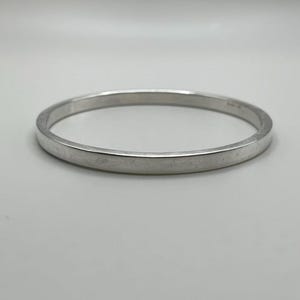 May include: A silver bangle bracelet with a flat, rectangular profile. The bracelet is a solid, continuous band of polished metal, reflecting light. The simple design suggests a minimalist aesthetic, suitable for everyday wear or as a layering piece.