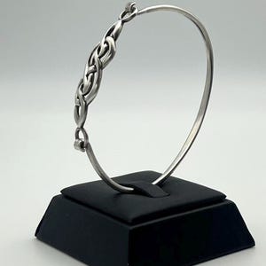 May include: A silver-toned metal bangle bracelet featuring a central decorative section with an intricate, interwoven knot-like pattern. The bracelet has a smooth, thin circular band that connects to the detailed centerpiece on both sides.