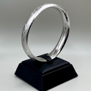 May include: A silver bangle bracelet with an engraved floral design. The hinged bracelet is displayed on a black stand against a neutral background. The bracelet's diameter is approximately 2.5 inches.