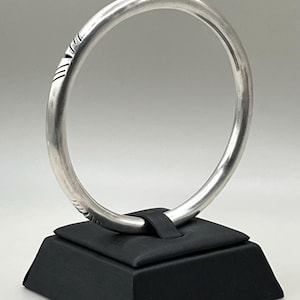 May include: A circular silver bangle with a smooth, polished finish and subtle engraved line patterns along the outer edge. The metal band features a consistent rounded profile and is displayed upright, showcasing its reflective surface and detailing.