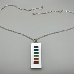 May include: A silver-toned metal necklace featuring a rectangular pendant. The pendant has a vertical row of six small, square-shaped inlays in a rainbow color sequence, including purple, blue, green, yellow, orange, and red, suspended on a chain.