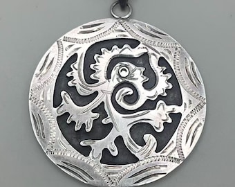May include: A round, silver pendant with an intricate, stylized design. The pendant features a central, dark-colored shape surrounded by a detailed, engraved border. The bail is a simple loop for a chain.