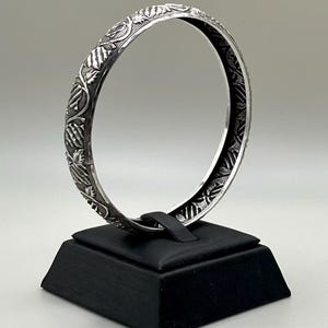 May include: A silver bangle bracelet with an intricate floral design. The bracelet is displayed on a black stand against a neutral background. The detailed pattern covers the entire surface of the bracelet.