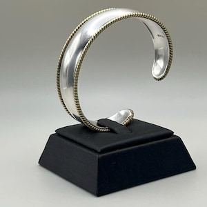 May include: A silver cuff bracelet featuring a smooth, polished center band flanked by two thin, twisted gold-tone rope borders. The bracelet has an open design, allowing it to be slipped onto the wrist, and displays a reflective metallic finish.