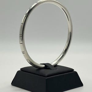 May include: A polished silver bangle bracelet with a smooth, rounded profile. The exterior surface features a series of evenly spaced, thin vertical etched lines that wrap around the circumference of the metal band, creating a subtle texture.