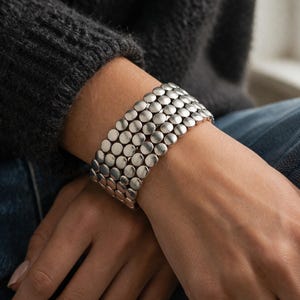 May include: A silver bracelet with a series of connected, circular, flat metal pieces. The bracelet is wide and wraps around the wrist. The bracelet is worn on a person's wrist, with the person wearing a dark gray sweater and blue jeans.