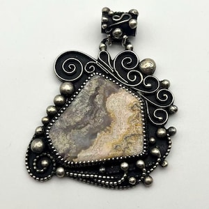 May include: A pendant featuring an irregular, multi-colored stone with swirling patterns of cream, gray, and brown. The stone is set in a dark, oxidized metal frame decorated with silver wire scrolls and small, round silver beads.