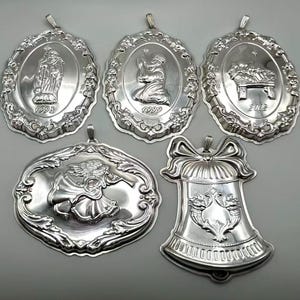 May include: Five silver Christmas ornaments with religious imagery. The ornaments include depictions of the Virgin Mary, an angel, and a nativity scene. Each ornament has a loop for hanging and is dated with the years 1998, 1999, and 2002.