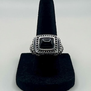 May include: A silver ring featuring a rectangular black stone set in the center. The metal band displays intricate beaded detailing around the stone setting and decorative scrollwork patterns along the sides of the band, creating a textured appearance.