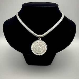 May include: A silver-toned necklace featuring a round pendant with an intricate embossed calendar design. The pendant is suspended from a thick, textured snake chain. The metal has a polished finish, and the circular centerpiece displays detailed radial patterns and central iconography.