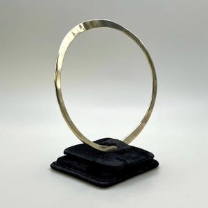 May include: A silver-toned, hammered metal bangle bracelet displayed on a black velvet jewelry stand. The bracelet has a slightly irregular, organic shape and a textured surface, reflecting light.