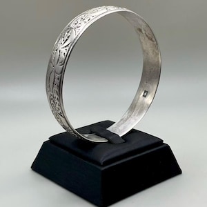 May include: A silver bangle bracelet with an engraved floral design. The bracelet is displayed on a black stand against a neutral background. The bangle is approximately 2.5 inches in diameter.