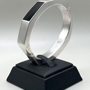 May include: A silver-toned hinged bangle bracelet featuring a geometric octagonal shape. The outer surface is accented with a smooth, rectangular black inlay panel. The bracelet has a polished metallic finish and a secure hinge mechanism.