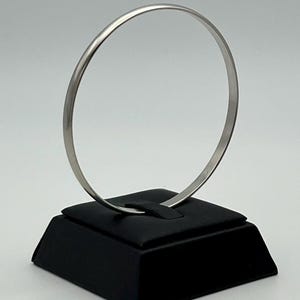 May include: A thin, circular silver-toned bangle bracelet with a smooth, polished finish. The bracelet is displayed upright, resting in a small, black, square-shaped jewelry stand designed to hold circular items securely in place.