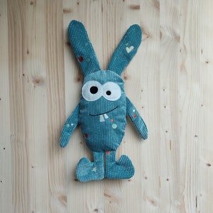 以下が含まれることがあります： 大きな目と歯を見せて笑っているモンスターの形をした、ターコイズブルーのぬいぐるみ。テクスチャのある生地で、白とオレンジの斑点で飾られています。