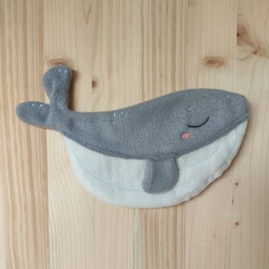 May include: A handmade grey and white felt whale toy with embroidered details. The whale has a smiling face with a pink mouth, closed eyes, and light blue stitching. The whale measures approximately 25 cm long.