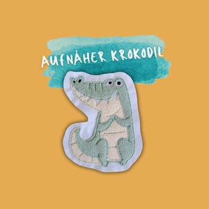 Könnte beinhalten: Ein grünes Filz-Krokodil-Appliké mit weißen Nähten und dem Text "AUFNÄHER KROKODIL" auf einem blauen Aquarellhintergrund.