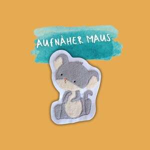 Könnte beinhalten: Eine graue Filz-Maus-Applikation mit dem Text "AUFNÄHER MAUS" auf einem blauen Aquarell-Hintergrund.