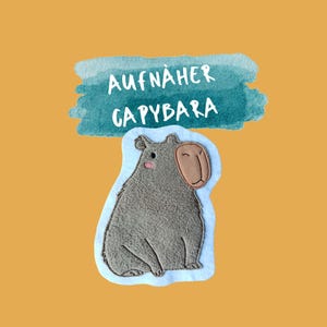 Könnte beinhalten: Ein grauer Capybara-Aufnäher mit hellblauem Rand, vor einem senfgelben Hintergrund. Der Aufnäher hat ein braunes Gesicht und rosa Wangen. Der Text "AUFNÄHER CAPYBARA" steht in Weiß über dem Capybara.