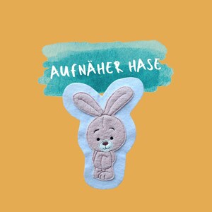 Könnte beinhalten: Ein Filz-Hasen-Applikation in Braun- und Beigetönen mit hellblauem Hintergrund. Der Hase hat große Ohren und ein lächelndes Gesicht. Der Text "AUFNÄHER HASE" befindet sich über dem Hasen auf einem türkisfarbenen Aquarellhintergrund, vor einem orangefarbenen Hintergrund.