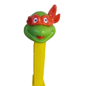 Op de afbeelding: Een groene Teenage Mutant Ninja Turtles-figuurkop met een rood masker op een gele plastic speelgoeddispenser.