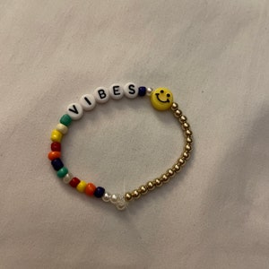 May include: A beaded bracelet with the word "VIBES" spelled out in white letters and a yellow smiley face charm. The bracelet is made with colorful beads, including red, orange, yellow, green, blue, and white.