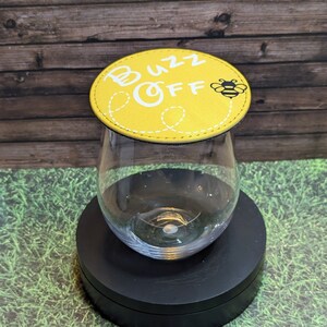Könnte beinhalten: Ein stieloses Weinglas mit einem gelben Untersetzer. Der Untersetzer trägt die weißen Worte "Buzz Off" und eine kleine Bienen-Grafik. Der runde Untersetzer hat einen genähten Rand. Das Glas ist leer.