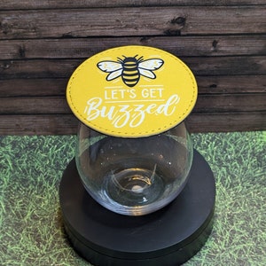 May include: A stemless wine glass with a yellow coaster featuring a bee illustration and the words "Let's Get Buzzed" in white. The coaster sits atop the glass, which is on a black stand.