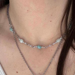 Puede incluir: Un collar de varias capas con una cadena plateada y pequeñas cuentas redondas de color azul claro y blanco. El collar se lleva alrededor del cuello.