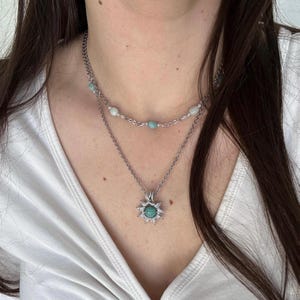 Può includere: Collana a strati con catena argentata. Lo strato superiore presenta piccole perline azzurre. Lo strato inferiore ha un ciondolo con una pietra turchese circondata da un motivo a raggiera. La collana è indossata su una maglietta bianca.