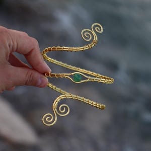Può includere: Un bracciale da braccio in filo metallico color oro con un design a spirale su entrambe le estremità e una singola pietra rotonda verde incastonata al centro della bobina. Il filo metallico è avvolto attorno alla struttura principale.