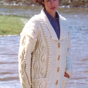 May include: Cream-colored, chunky knit cardigan with a leaf pattern and wooden buttons. The long-sleeved sweater has two front pockets and a shawl collar. The woman is wearing a denim shirt underneath.