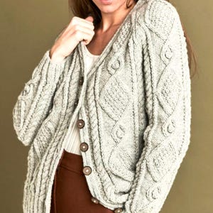 May include: A light grey, button-down cardigan with a cable knit pattern. The sweater has a V-neck and is made of a soft, textured material. The buttons are dark brown. The cardigan is worn over a white top and brown trousers.
