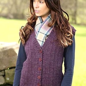 Puede incluir: Una mujer con un chaleco morado de punto con botones, sobre una camisa azul de manga larga y una bufanda a cuadros. También lleva una falda a cuadros. El fondo es una escena borrosa al aire libre.