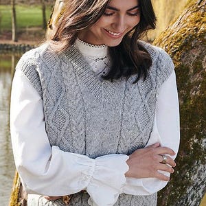 Puede incluir: Un chaleco de punto gris con cuello en V, tejido con trenzas, usado sobre una camisa blanca de manga larga con puños con volantes. El chaleco tiene un patrón texturizado. El estilo es clásico y adecuado para combinar.