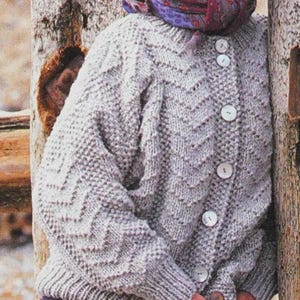 May include: A light gray, button-up knit cardigan with a chevron pattern, paired with purple plaid pants. The outfit includes a patterned scarf and a brown leather bag with a strap.