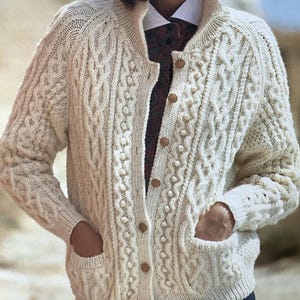 May include: Cream-colored cable-knit cardigan with a round collar, wooden buttons, and two front pockets. The sweater features intricate cable patterns and bobble stitch details. A classic, warm, and textured knitwear piece.