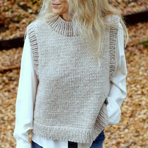 May include: A light beige, knitted vest with a high, ribbed collar and ribbed edges. The sleeveless sweater is worn over a white collared shirt. The vest has a textured, chunky knit design.