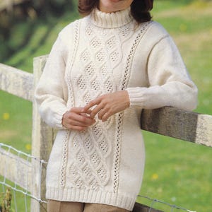 May include: Cream-colored turtleneck sweater with intricate cable-knit and openwork detailing. The long-sleeved sweater features a textured pattern down the front and along the sides. The model is leaning on a wooden fence.