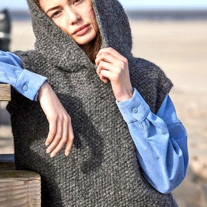 Könnte beinhalten: Eine graue Kapuzenweste mit strukturiertem Strickdesign. Die Weste wird über einem hellblauen Button-Down-Hemd getragen. Das Model lehnt an einer Holzkonstruktion, mit einem Strand im Hintergrund.