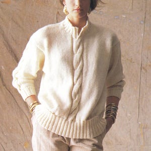 Könnte beinhalten: Cremefarbener Zopfstrickpullover mit hohem Kragen und zentralem Zopfmuster. Der Pullover hat lange Ärmel und einen gerippten Saum und Bündchen. Das Model trägt goldene Armbänder und Ohrringe. Der Pullover wird mit Cordhosen kombiniert.