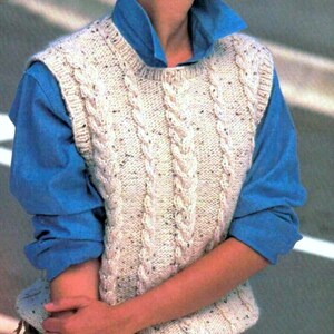 Può includere: Un gilet color crema a maglia a trecce con macchie di colore scuro. Il gilet è senza maniche e ha uno scollo tondo. Il gilet è indossato sopra una camicia blu con colletto. Il design a maglia a trecce è uno stile classico e senza tempo.