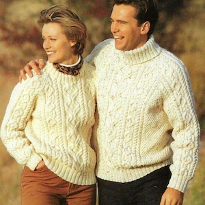 May include: Two people wearing cream-coloured cable-knit jumpers. The jumpers have a textured pattern and long sleeves. One person is wearing brown trousers, and the other is wearing black trousers. The background is blurred.