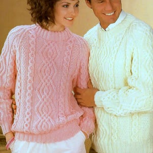 Puede incluir: Un suéter de punto trenzado rosa claro con cuello alto y patrones intrincados. El suéter tiene mangas largas y un dobladillo acanalado. Una persona lleva el suéter. Otra persona lleva un suéter de punto trenzado color crema.