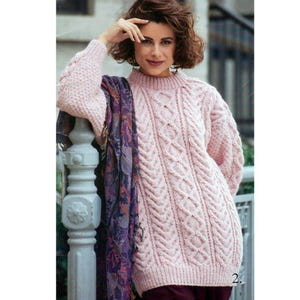May include: A pale pink cable-knit jumper with a high neckline and long sleeves. The jumper has a textured pattern. A patterned scarf in purple and blue is draped over the shoulder. The garment is a warm and cosy knit.