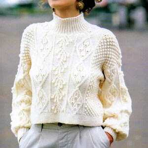 May include: Cream-colored, textured knit sweater with a high, ribbed collar. The sweater features intricate cable-knit and raised floral patterns across the chest and sleeves. The sleeves are long and the cuffs are ribbed.