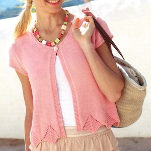 Könnte beinhalten: Hellrosa, kurzärmliger Cardigan mit gewelltem Saum. Der offene Pullover hat einen einzelnen Knopfverschluss und wird mit einem weißen Oberteil und einem beigen Rock kombiniert. Eine bunte Halskette und eine Strohtasche vervollständigen den Look.