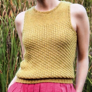 May include: Mustard yellow knitted tank top with a textured pattern. The sleeveless top has a round neckline and a ribbed hem. The top is paired with a pink skirt. The background features green foliage.