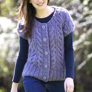 Op de afbeelding: Een lila gekleurd vest met korte mouwen, een kabelgebreid patroon en grote, ronde knopen. Het vest wordt gedragen over een donkerblauwe longsleeve en gecombineerd met een blauwe jeans. De algehele stijl is casual en comfortabel.